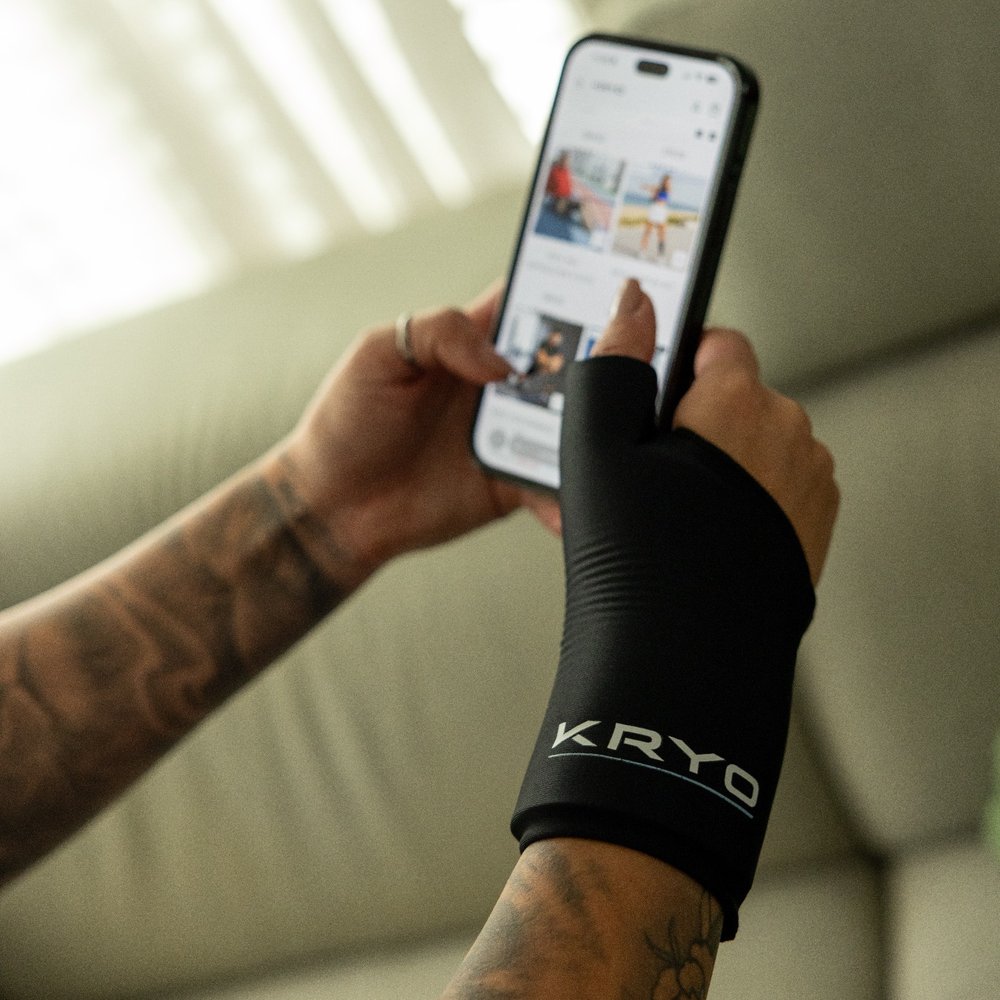 Woman wearing the KRYO Wrist Sleeve while using her smartphone indoors, with soft natural light filtering through white curtains.