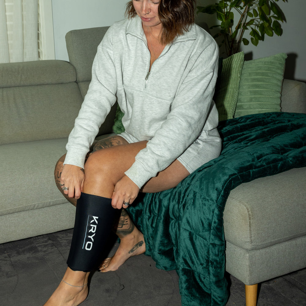 Woman sitting on a couch putting on a KRYO CORE knee and calf ice compression sleeve for at-home recovery.