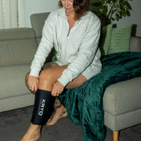 Woman sitting on a couch putting on a KRYO CORE knee and calf ice compression sleeve for at-home recovery.