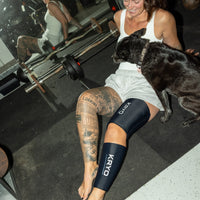 Woman relaxing on a gym floor with her dog while wearing KRYO MAX thigh and CORE knee and calf ice compression sleeves for post-workout recovery.