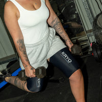 Woman performing dumbbell lunges in a gym while wearing two KRYO MAX thigh ice compression sleeves for post-workout recovery.